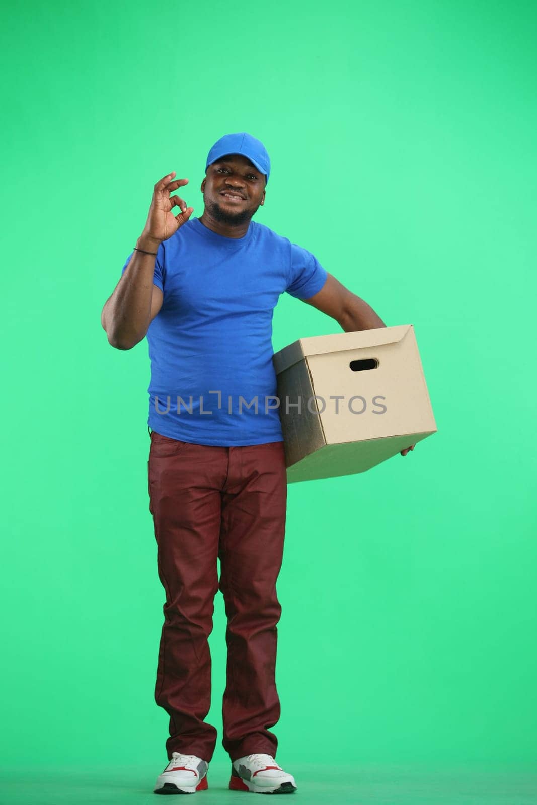 The deliveryman, in full height, on a green background, with a box, shows the ok sign.
