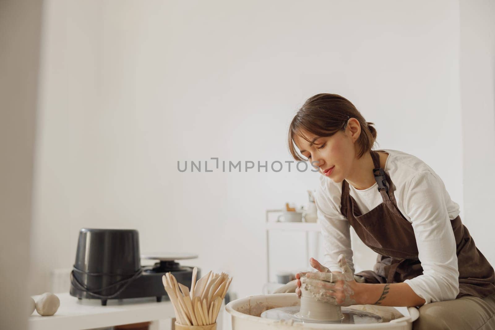Concentrated female artisan in apron sitting on bench with pottery wheel and making clay pot