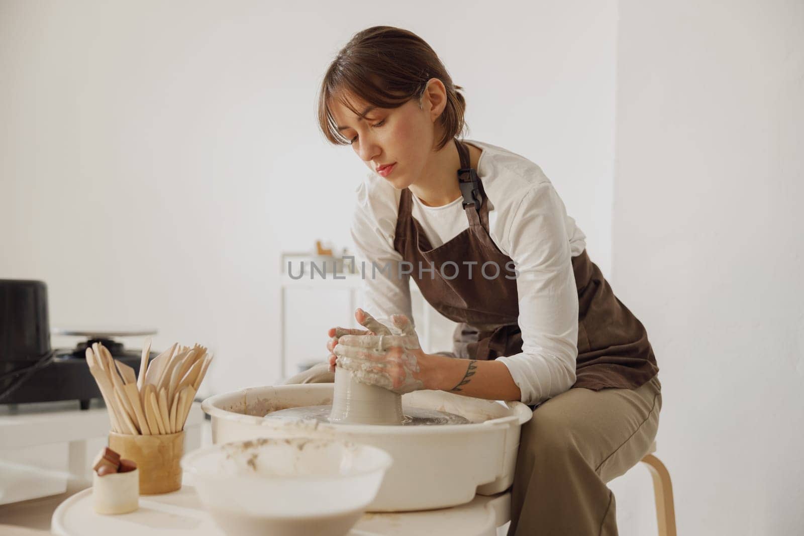 Concentrated female artisan in apron sitting on bench with pottery wheel and making clay pot by Yaroslav_astakhov