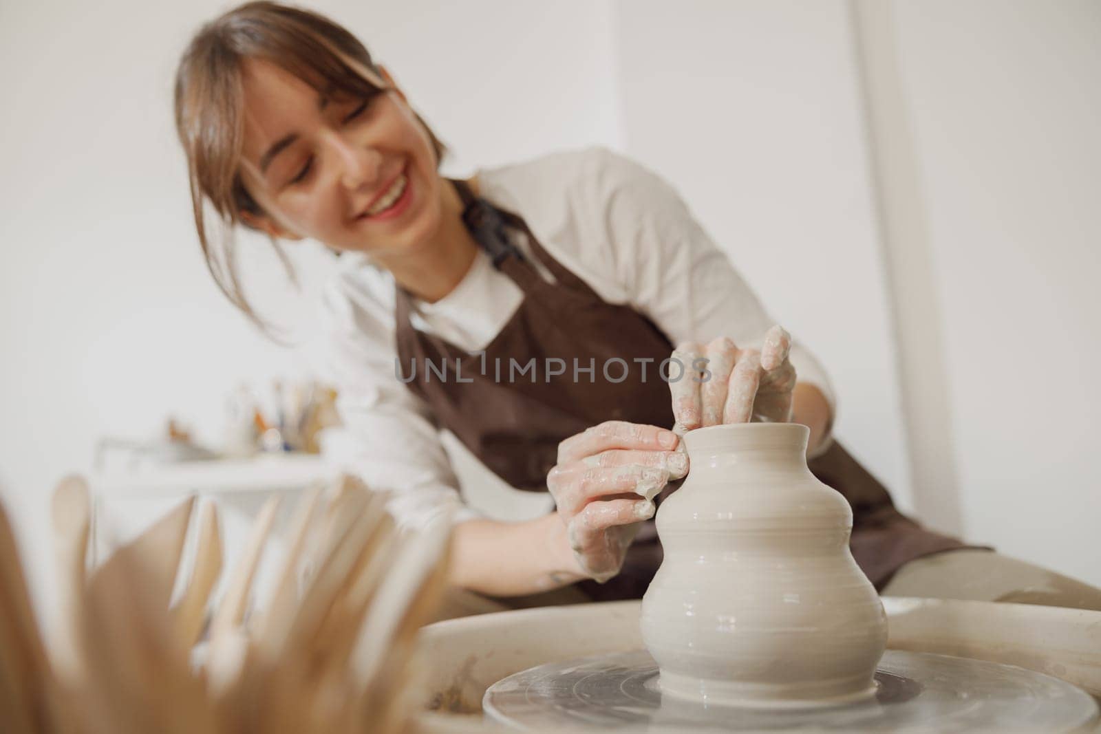 Smiling female artisan in apron sitting on bench with pottery wheel and making clay pot by Yaroslav_astakhov