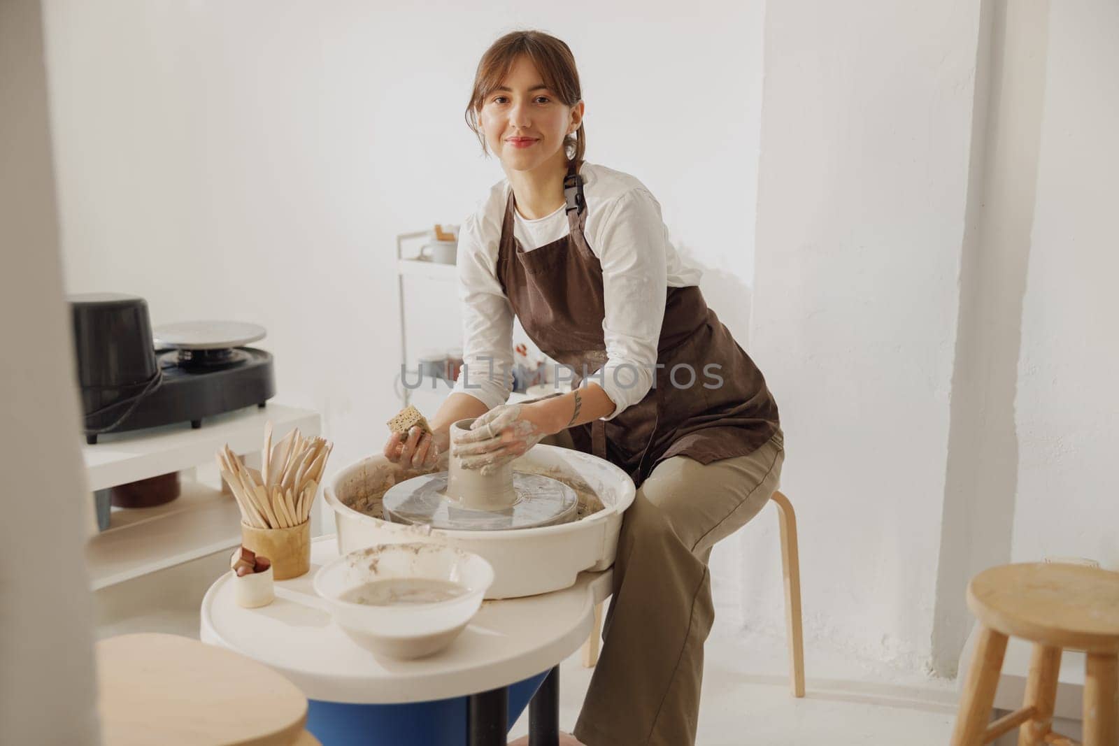 Smiling female artisan in apron sitting on bench with pottery wheel and making clay pot by Yaroslav_astakhov