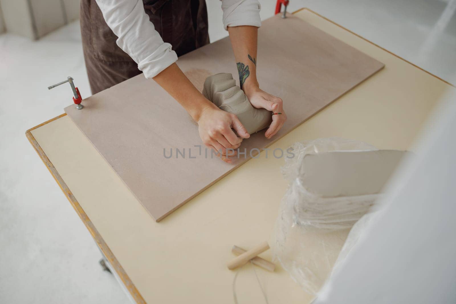 Close up of female potter in apron kneads piece of clay with her hands on table in studio by Yaroslav_astakhov