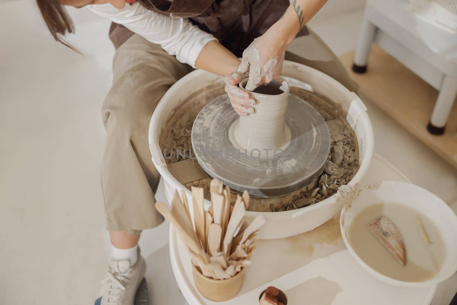 Female potter in apron making shape of clay vase on spinning pottery tool in ceramic workshop by Yaroslav_astakhov