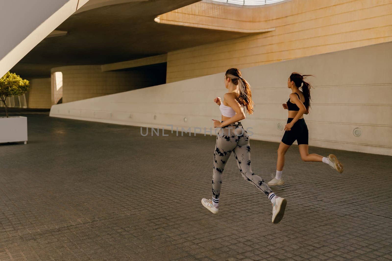 Two young women in sportswear are running on modern buildings background. Active lifestyle concept by Yaroslav_astakhov