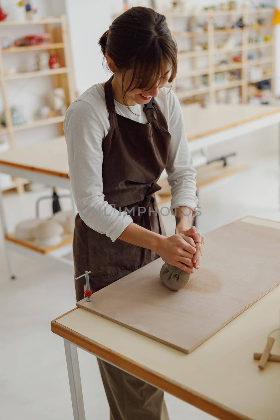 Professional female potter in apron kneads piece of clay with her hands on table in studio