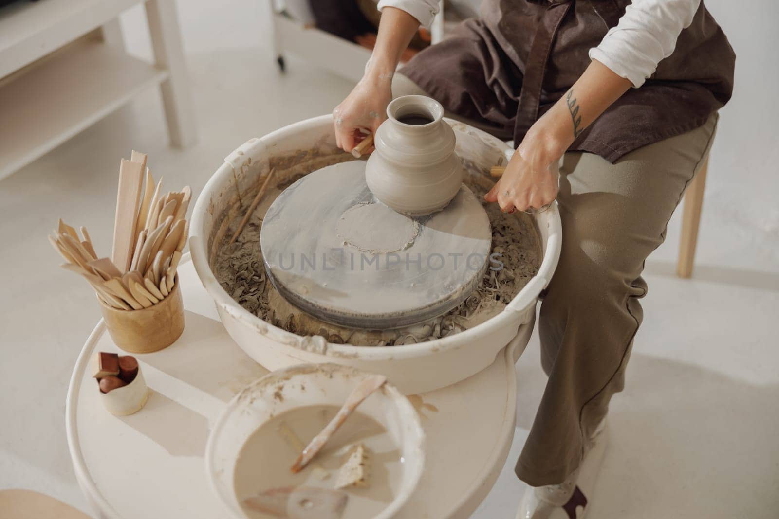 Female potter in apron making shape of clay vase on spinning pottery tool in ceramic workshop by Yaroslav_astakhov