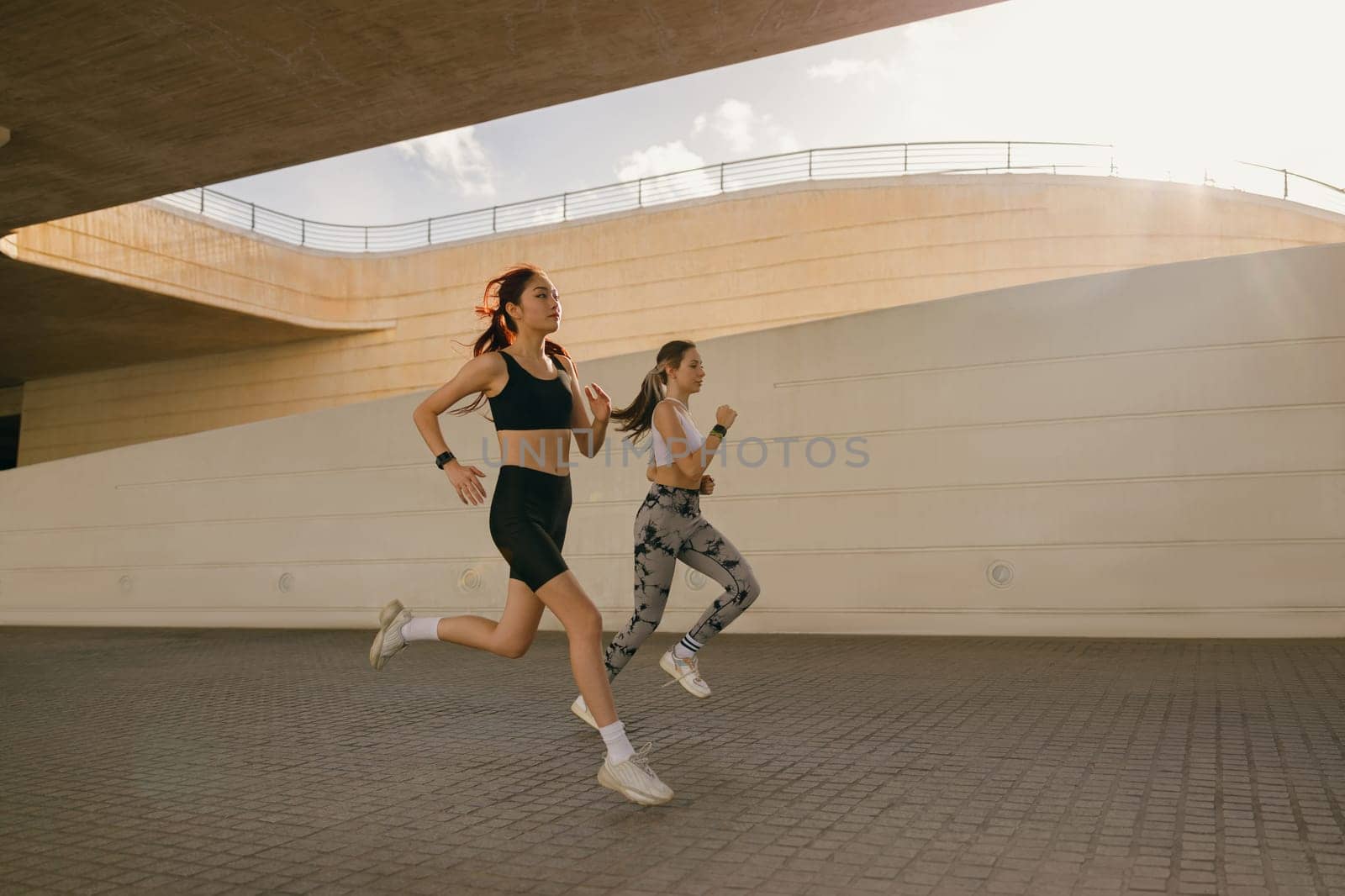 Two active women athlete running on sunny morning side by side on modern buildings background