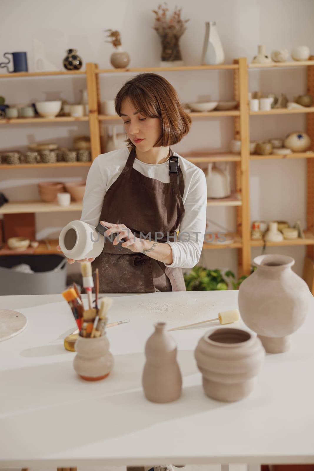 Young female potter holding unfired clay vase while standing in pottery studio. High quality photo