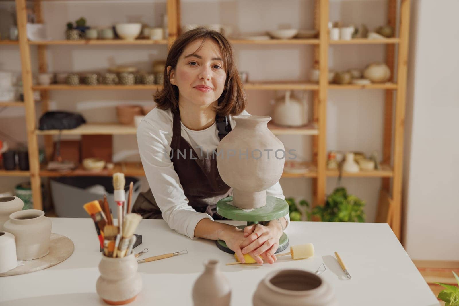 Portrait of young female potter in apron with mug looking at camera while posing in workshop by Yaroslav_astakhov