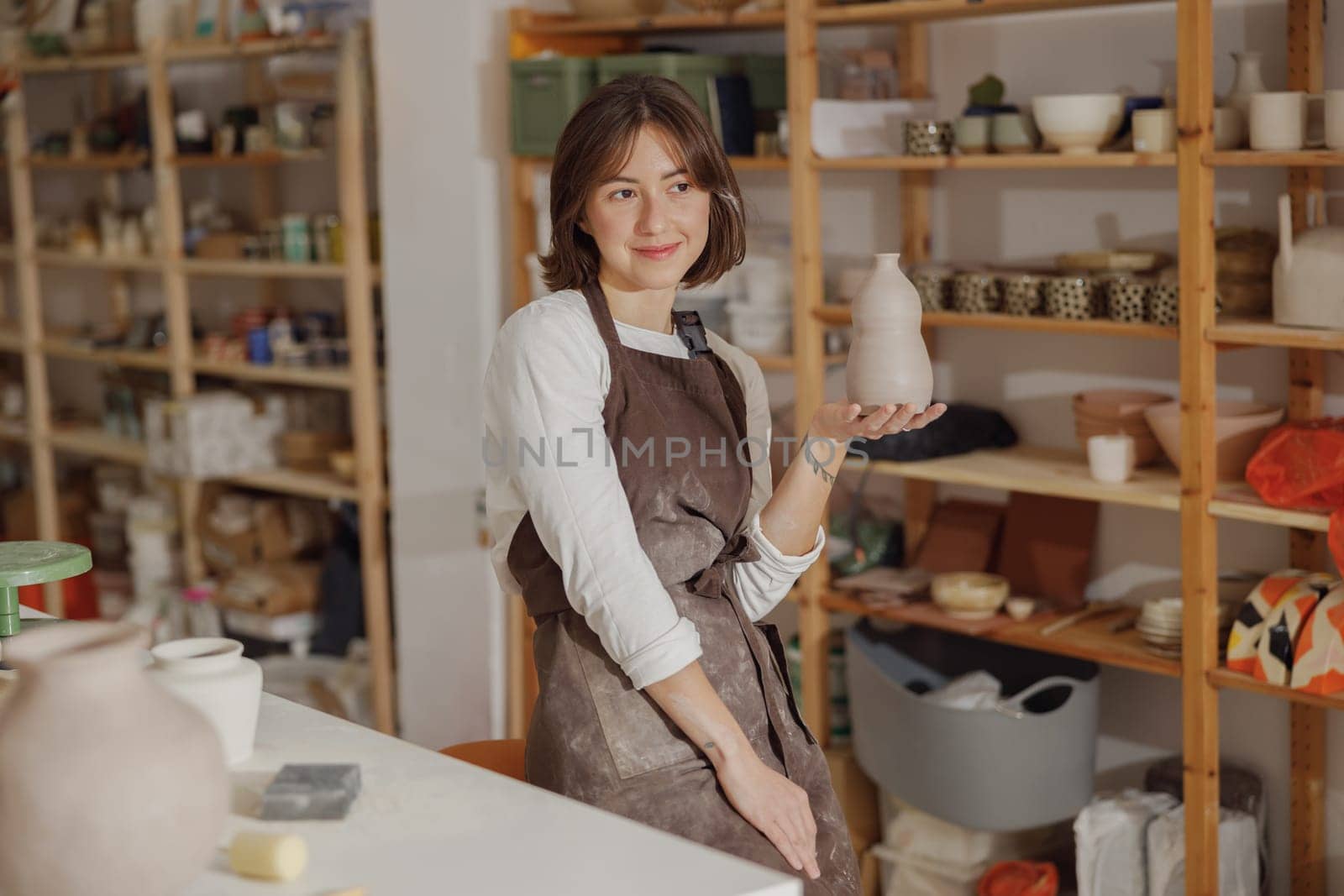 Portrait of young female potter in apron with mug looks away while posing in workshop by Yaroslav_astakhov