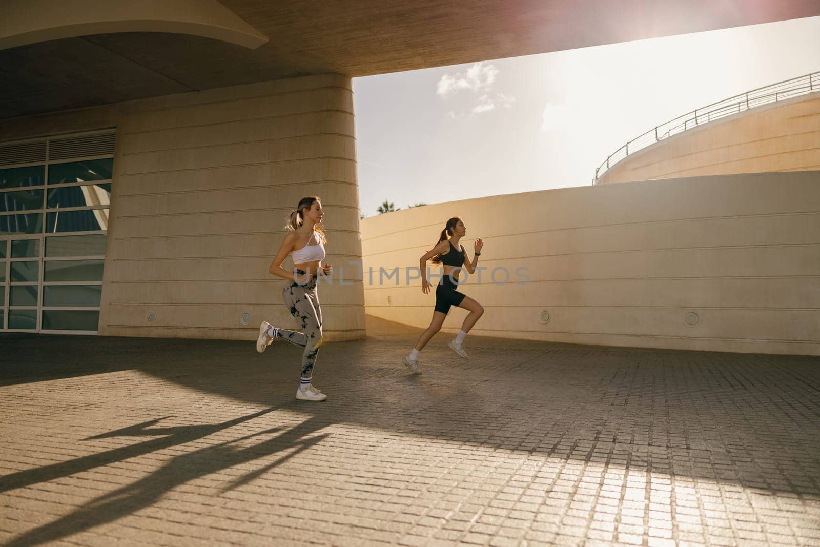 Two young women in sportswear are running on modern buildings background. Active lifestyle concept by Yaroslav_astakhov