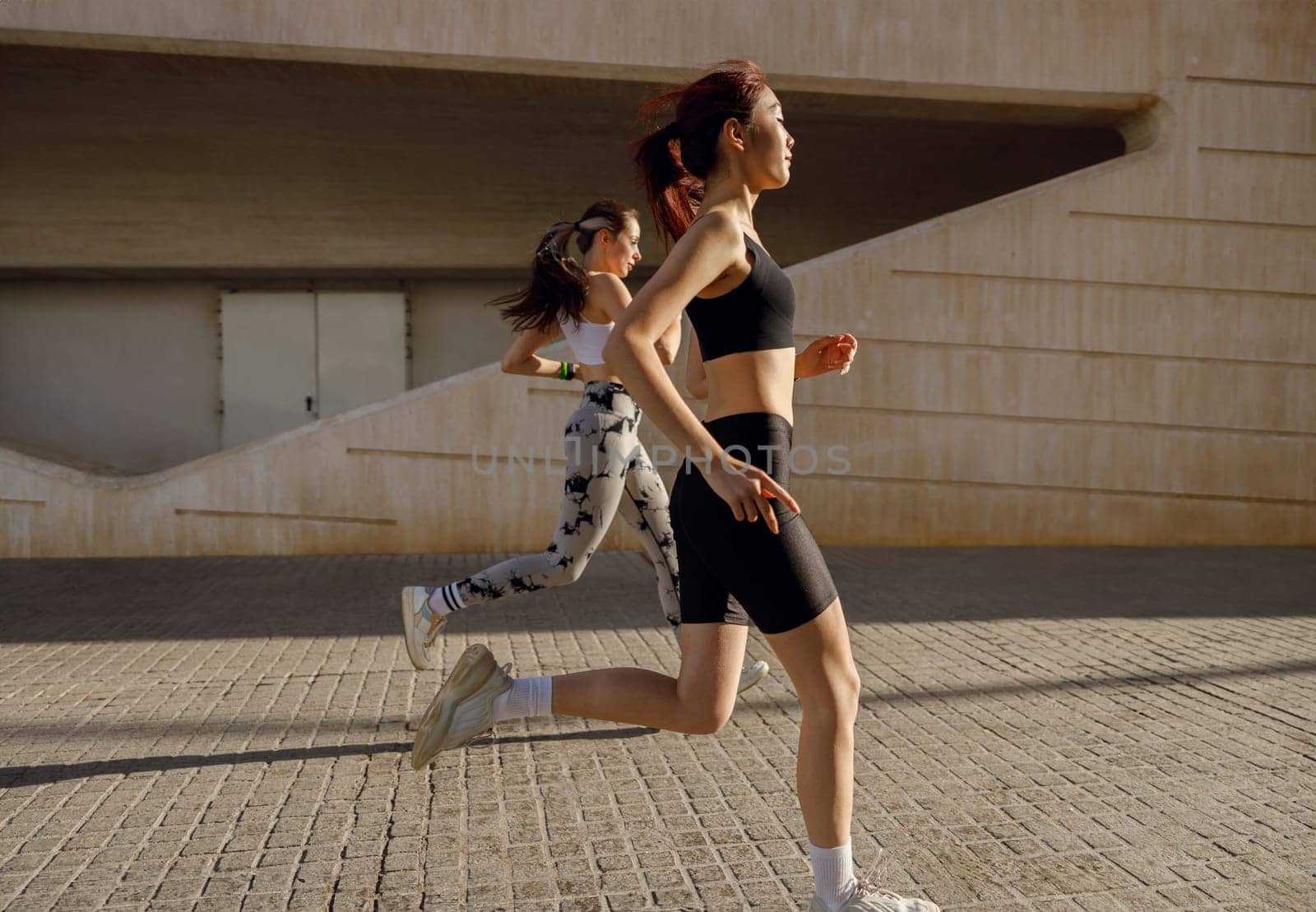 Active female friends athlete are running on sunny morning on modern buildings background by Yaroslav_astakhov