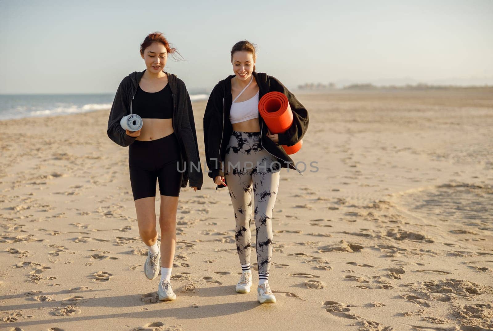 Two young fit women walking on the beach with their yoga mats after doing morning gym by Yaroslav_astakhov