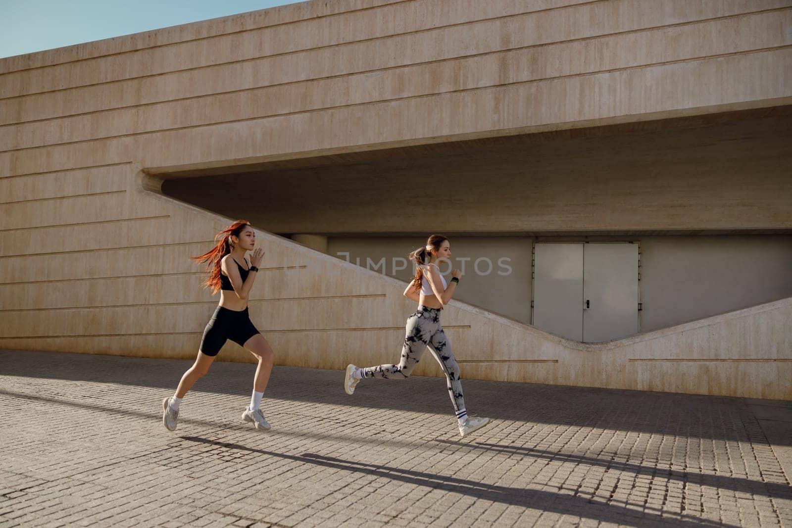 Two active women athlete running on sunny morning side by side on modern buildings background by Yaroslav_astakhov
