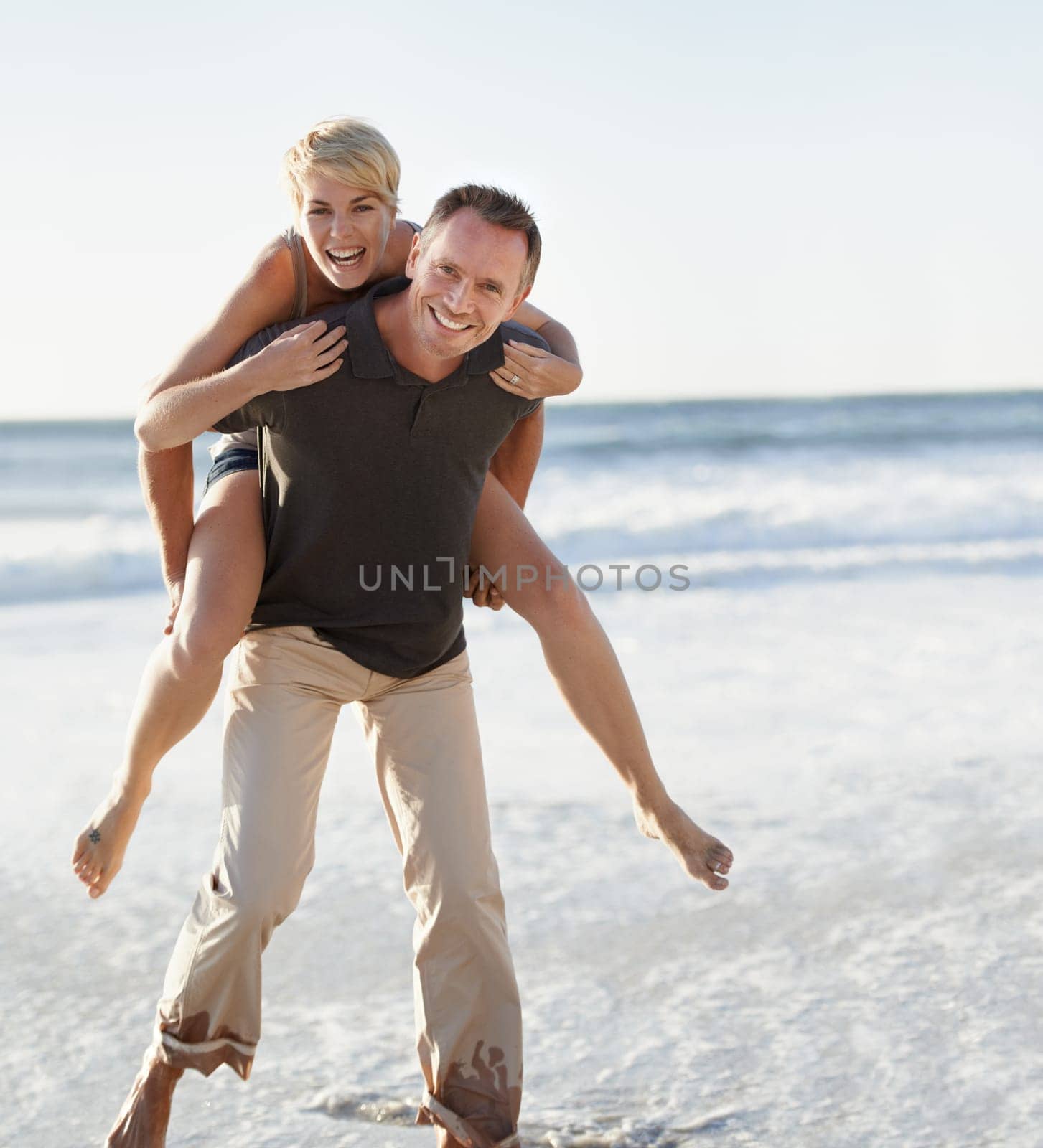 Couple, portrait and happy on beach with piggyback for bonding, honeymoon date and weekend holiday. Face, man and woman with embrace, smile and love in water by sea for vacation, travel and adventure by YuriArcurs