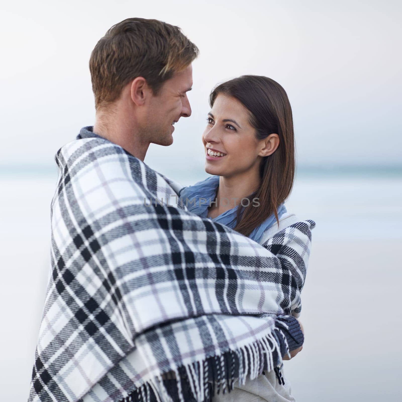 Couple, hug and smile at beach with blanket for vacation, love and care on date at sunset by ocean. Man, woman and embrace with break, bonding and travel for memory on holiday by sea in Australia.