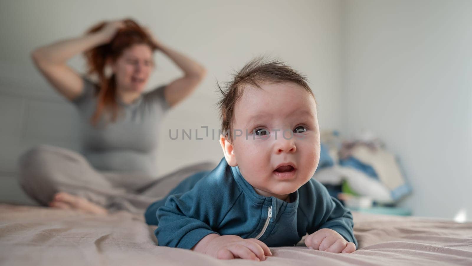 A three-month-old boy lies on his stomach on the bed and his mother sits behind him and cries. Postpartum depression