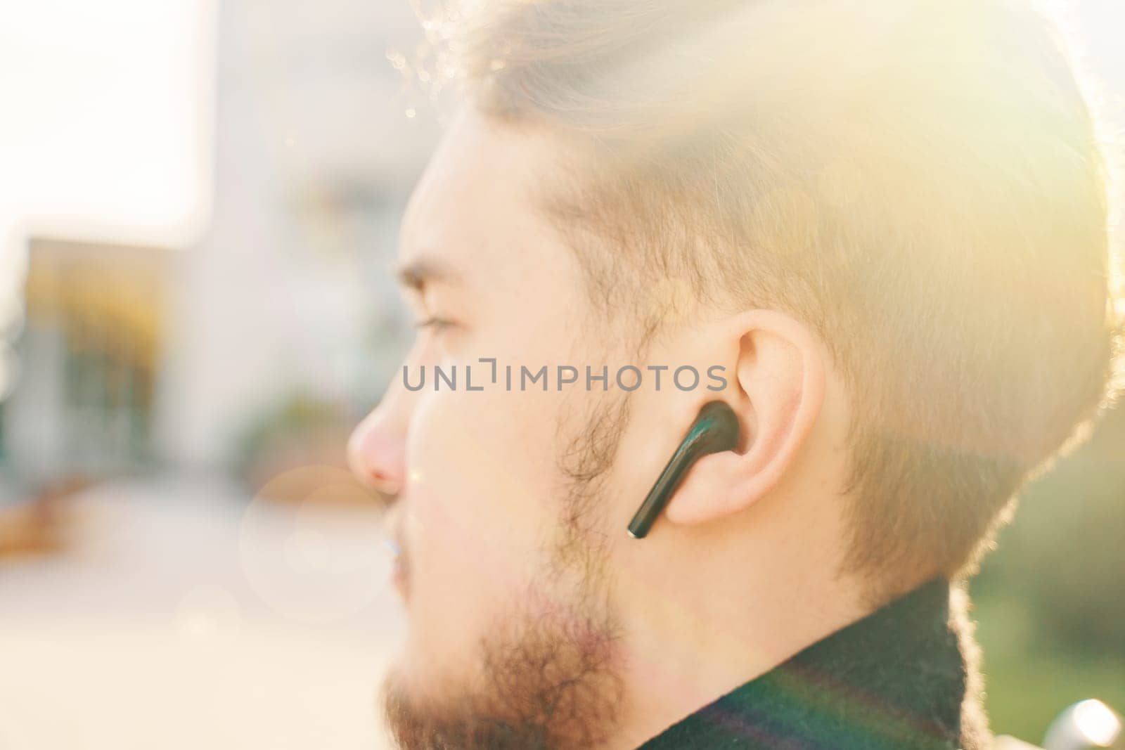 Young man in a coat and wireless headphones sits on a Park bench