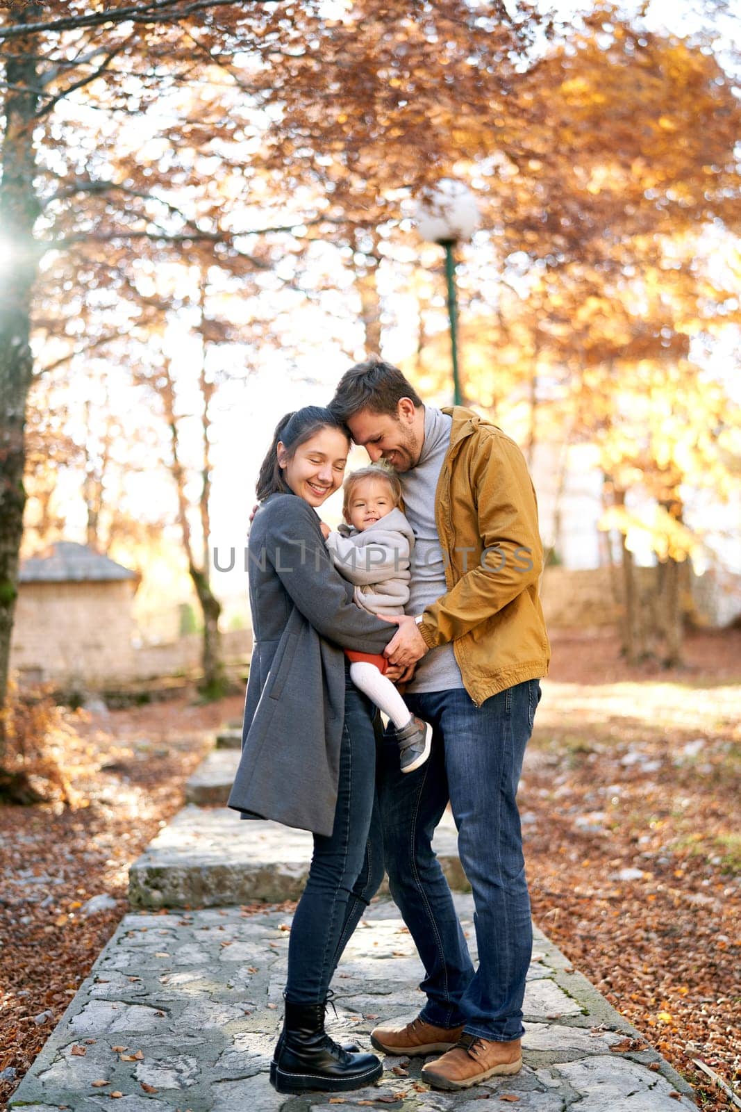 Smiling dad and mom touching foreheads while standing with a little girl in her arms in the autumn forest. High quality photo