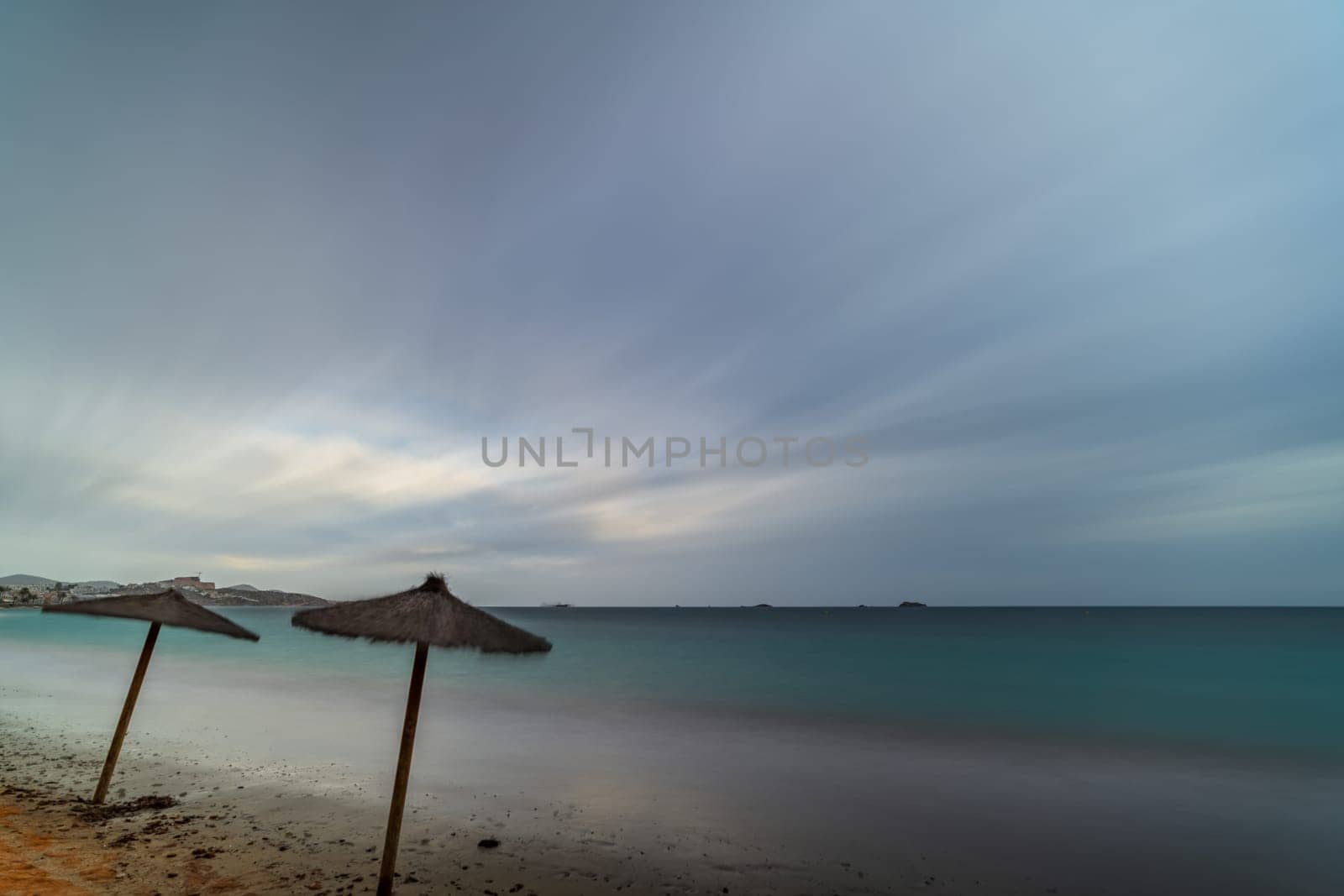 Stormy day long exposure shot of an empty white sandy beach with straw umbrellas, creating a dark, mysterious mood.