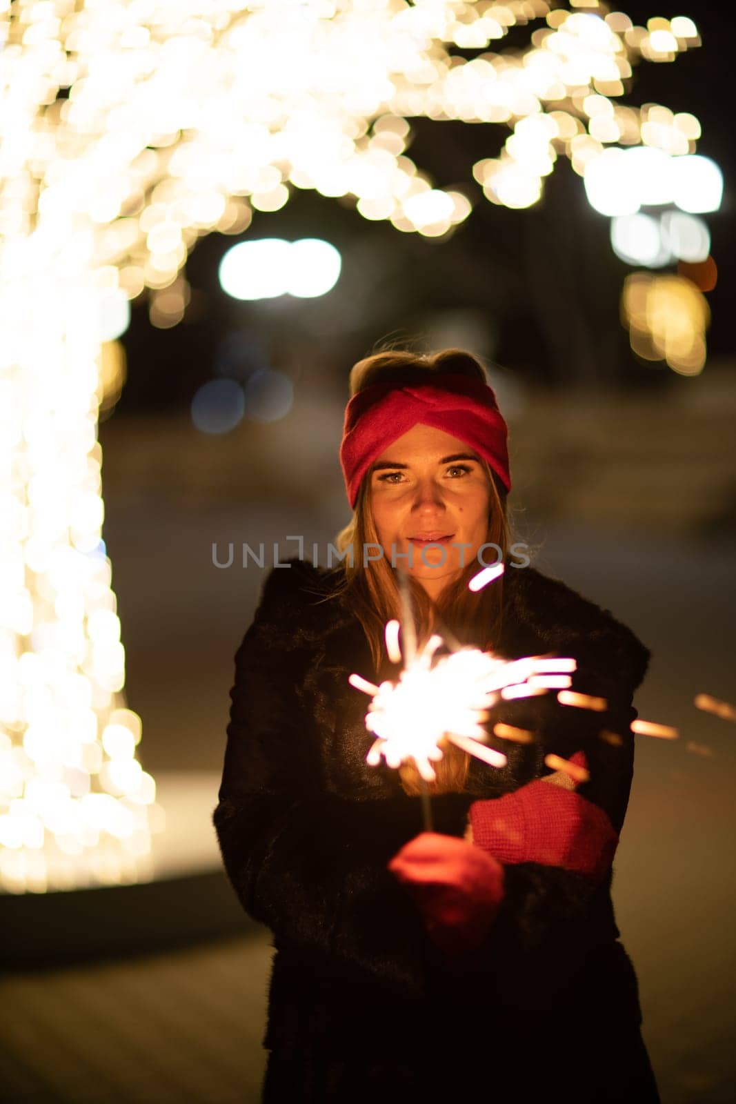 Woman holding sparkler night while celebrating Christmas outside. Dressed in a fur coat and a red headband. Blurred christmas decorations in the background. Selective focus.