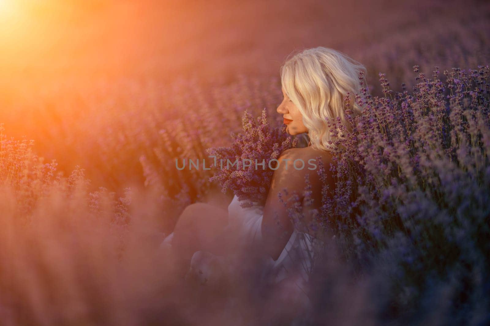 Blonde woman poses in lavender field at sunset. Happy woman in white dress holds lavender bouquet. Aromatherapy concept, lavender oil, photo session in lavender.