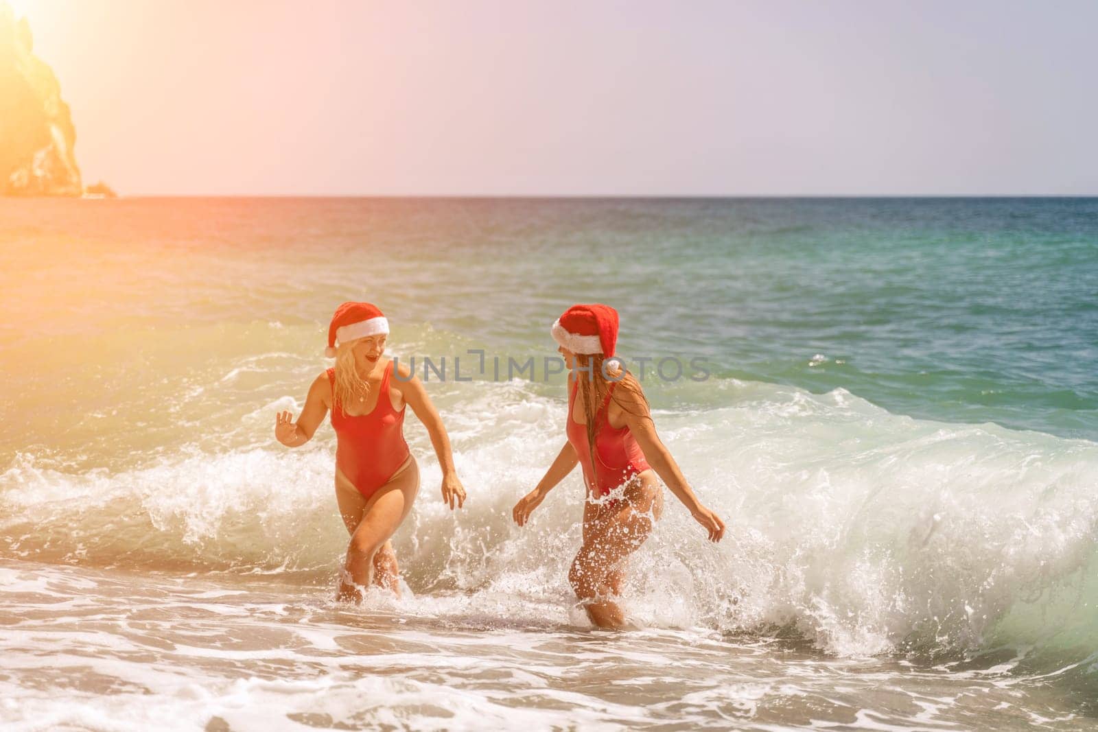 Women Santa hats ocean play. Seaside, beach daytime, enjoying beach fun. Two women in red swimsuits and Santa hats are enjoying themselves in the ocean waves