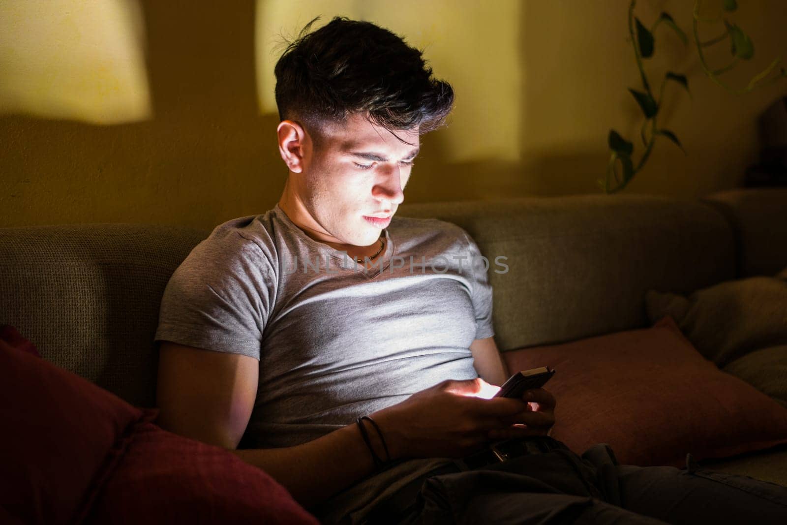 A man is seated on a couch, engrossed in his cell phone screen. He appears focused as he scrolls and interacts with the device, possibly texting or browsing. The background shows a typical living room setting.