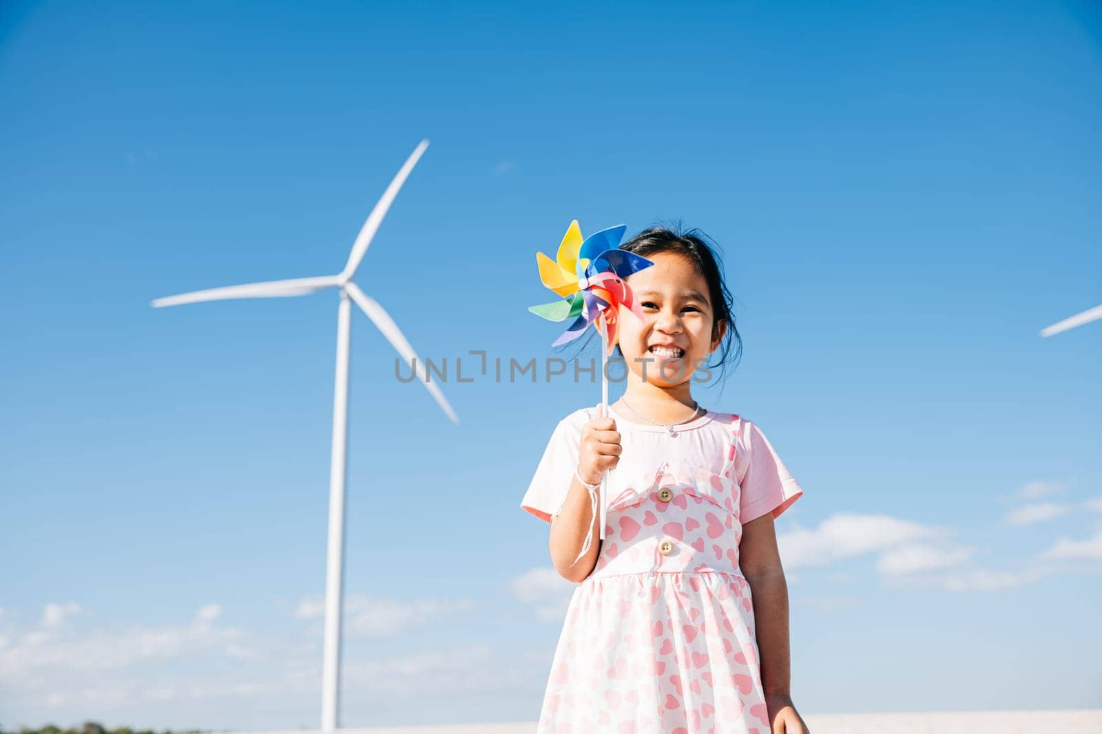 Little girl's playful joy near windmills, running with pinwheels. Illustrates wind energy education promoting clean electricity and sustainable industry in a picturesque setting.