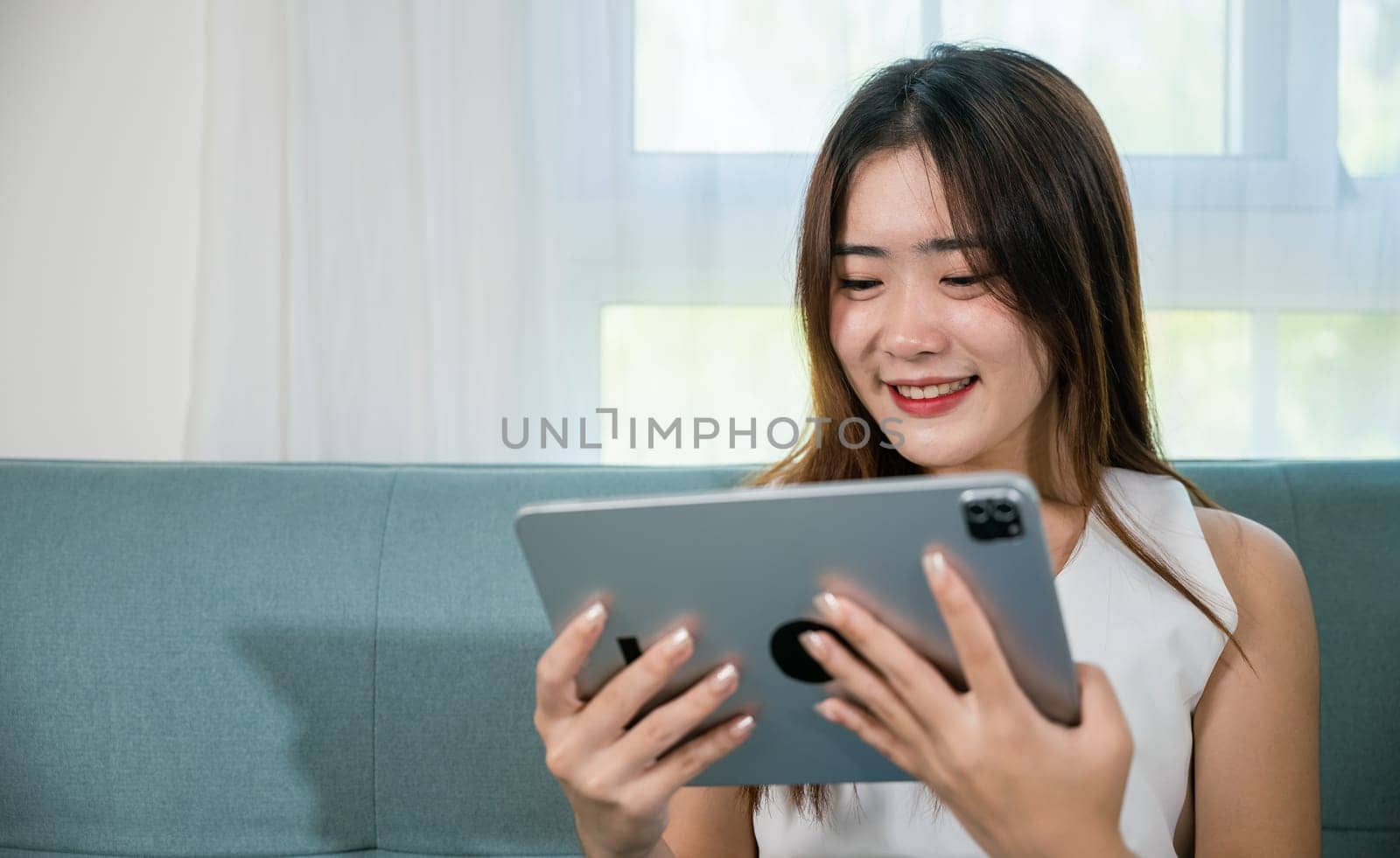 Happy Asian woman playing smart tablet at home while relaxing on sofa living room by Sorapop