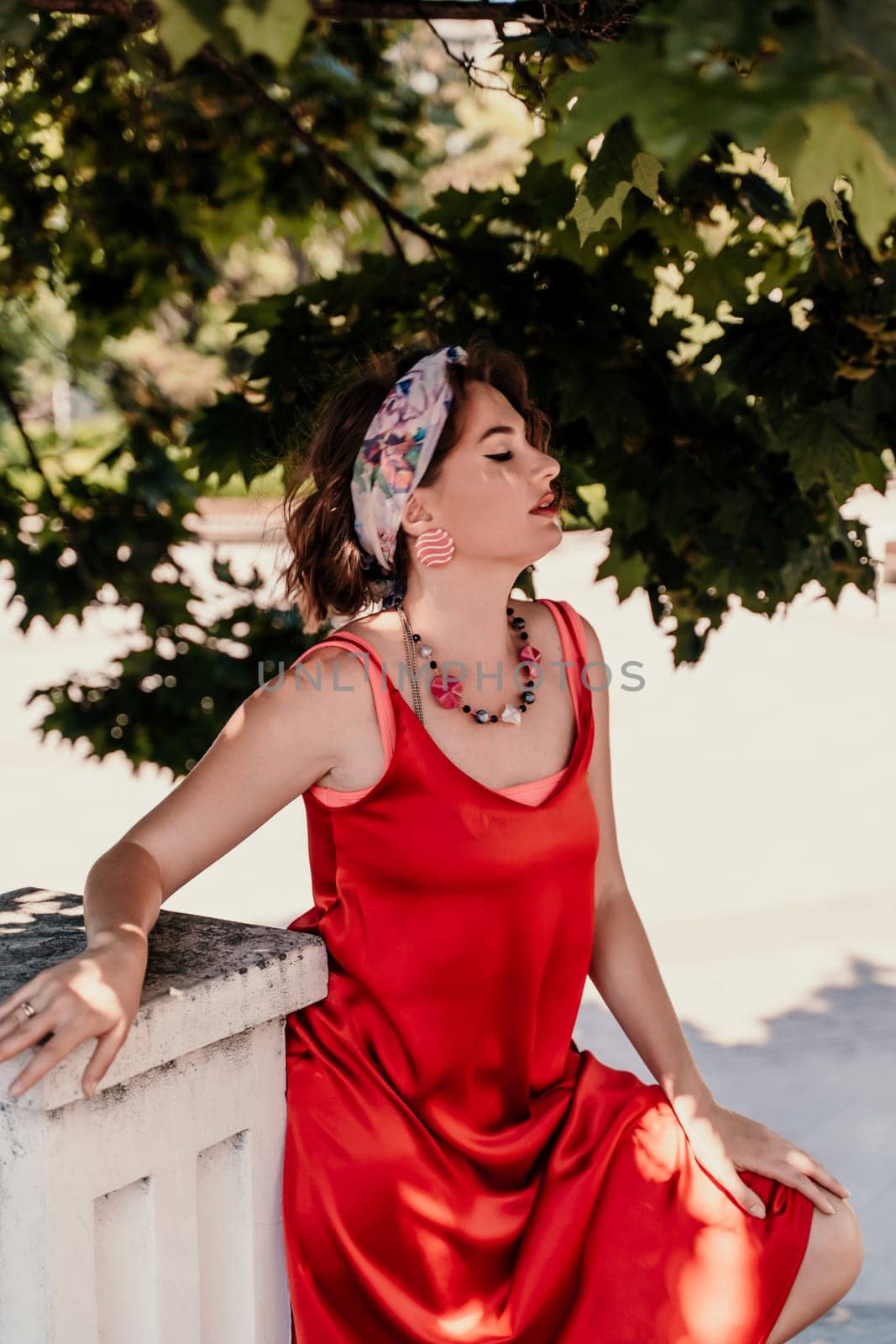 woman in a red silk dress and a bandage on her head smiles against the background of the leaves of a tree. She is leaning on the coop and looking into the camera. Vertical photo