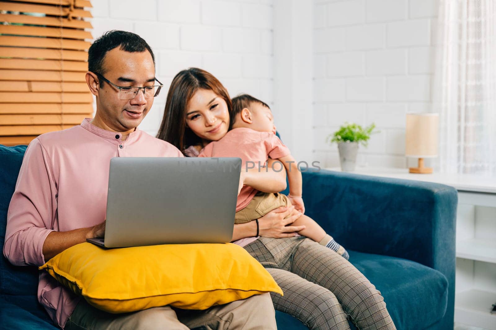 A businesswoman carries her sleeping daughter while her husband works on a laptop by Sorapop