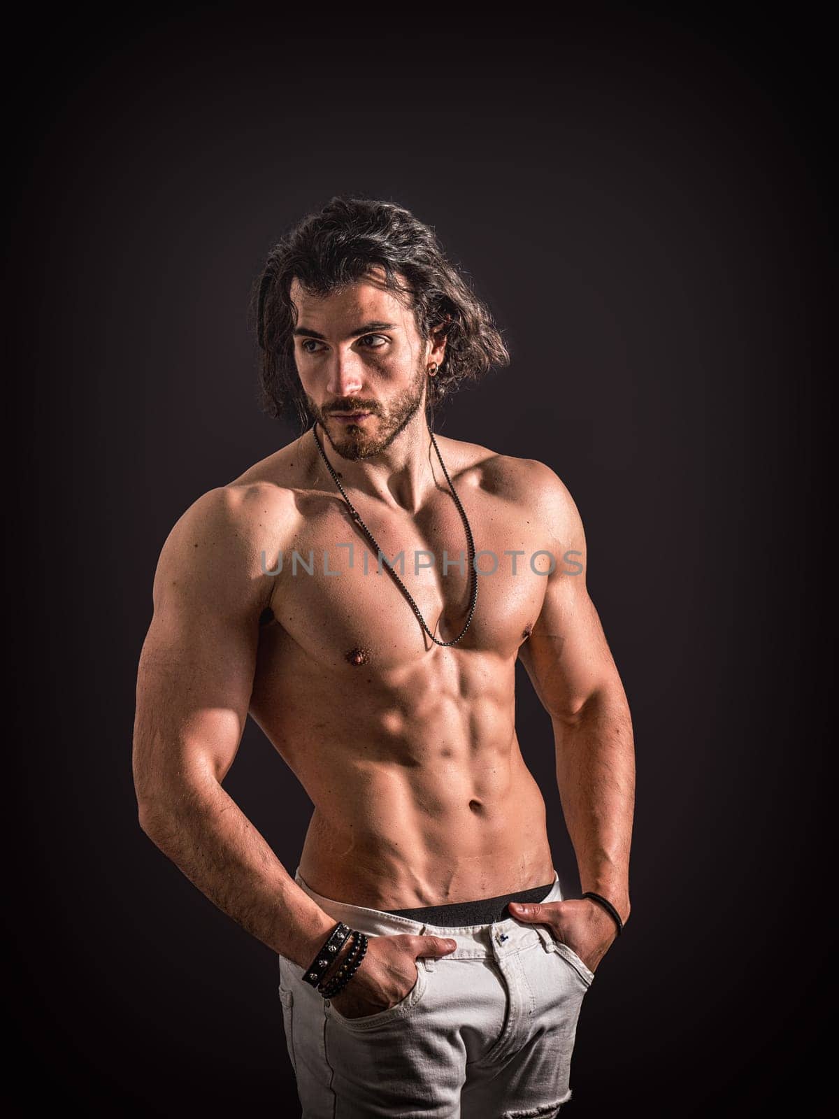 A man with long hair is standing shirtless, striking a pose for a photo. His toned physique is highlighted as he confidently gazes at the camera.