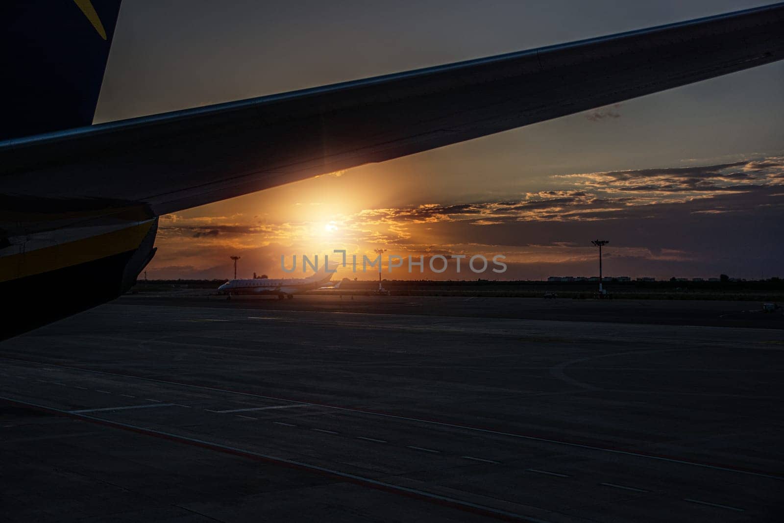 The image captures the sun gradually setting in the horizon, casting a warm glow behind the wing of a commercial airplane in flight. The silhouette of the aircraft contrasts against the vibrant colors of the sky, creating a striking composition.