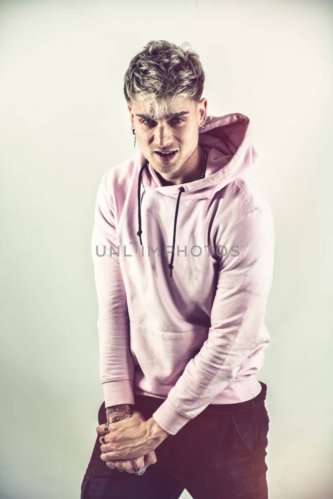 A man wearing a pink hoodie stands with his hands clasped together in front of him. The background is neutral, allowing the focus to be on the man and his clothing.