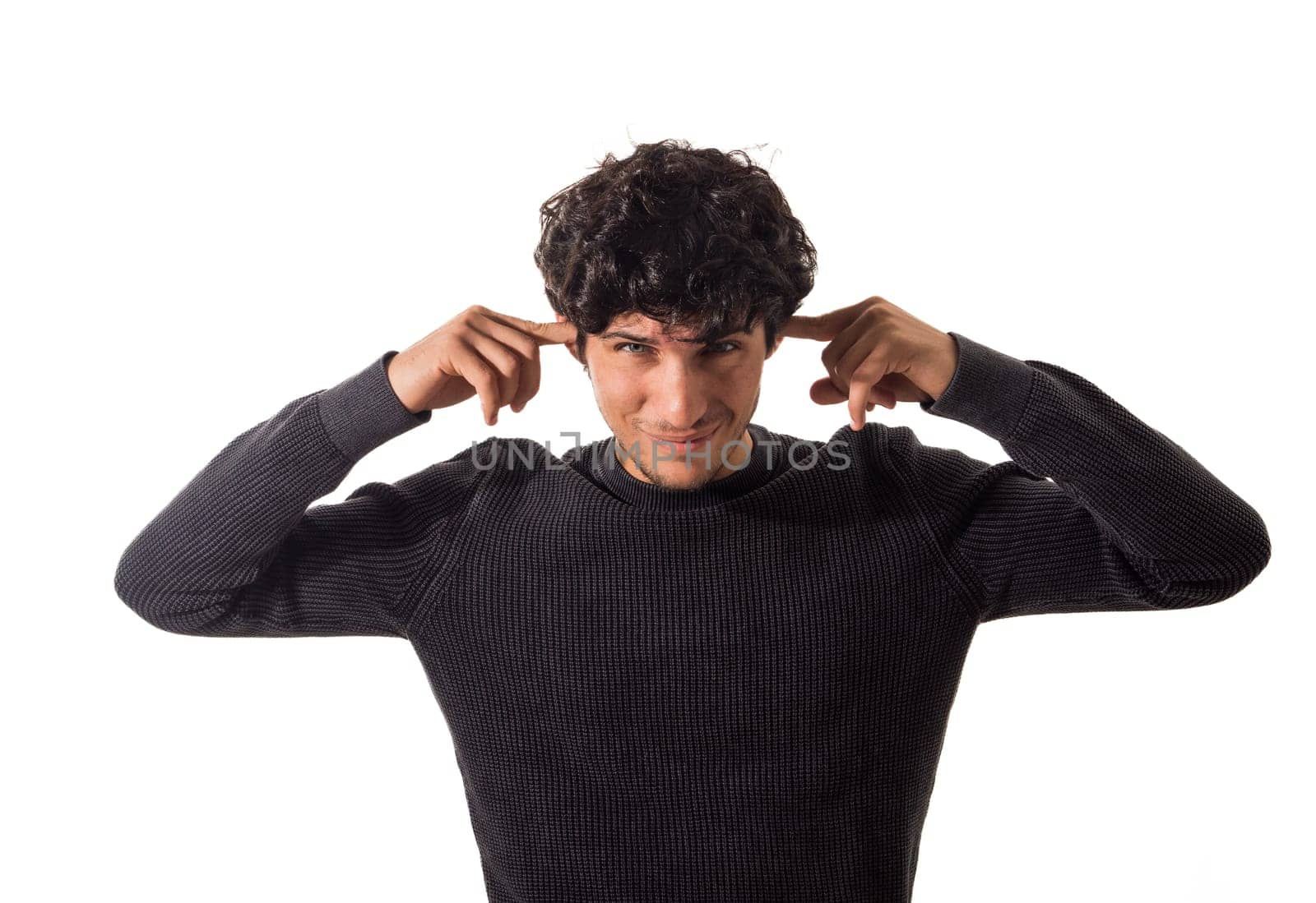 Too much noise: attractive young man covering his ears. A young man with curly hair is seen wearing a black sweater. He is standing against a neutral background, with the focus on his clothing and hair.