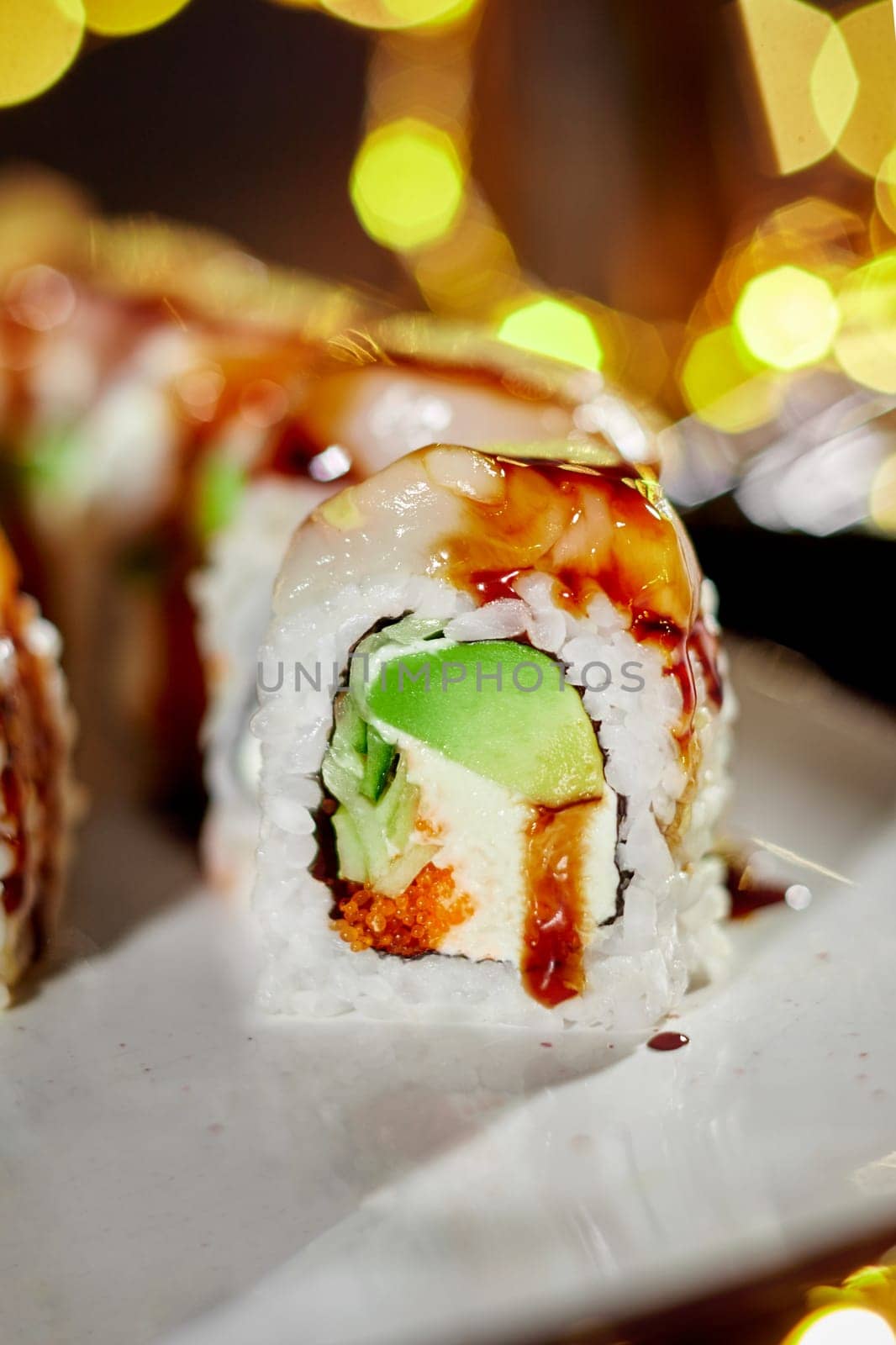 Closeup of appetizing sushi roll with sea scallop, cream cheese, masago roe, cucumbers and avocado seasoned with spicy mayo an unagi sauce served against warm festive lights