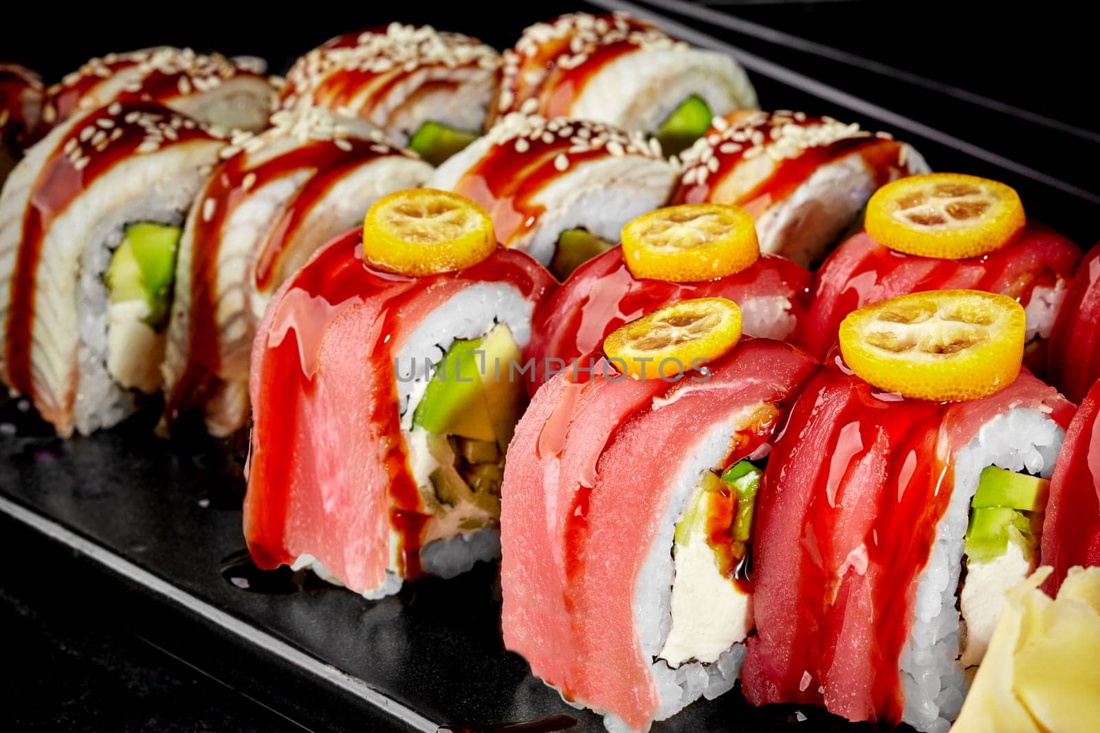 Closeup of delicious fresh sushi rolls filled with cream cheese, avocado and cucumber topped with tuna and eel seasoned with unagi sauce garnished with slices of kumquat and sesame on black background