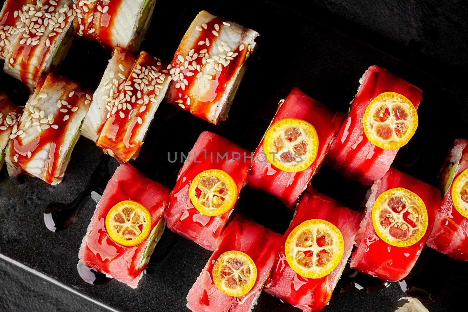 Top view of diverse sushi platter featuring two varieties, topped with tuna and kumquat slices and other with eel sprinkled of sesame seeds, drizzled with spicy unagi sauce on black background