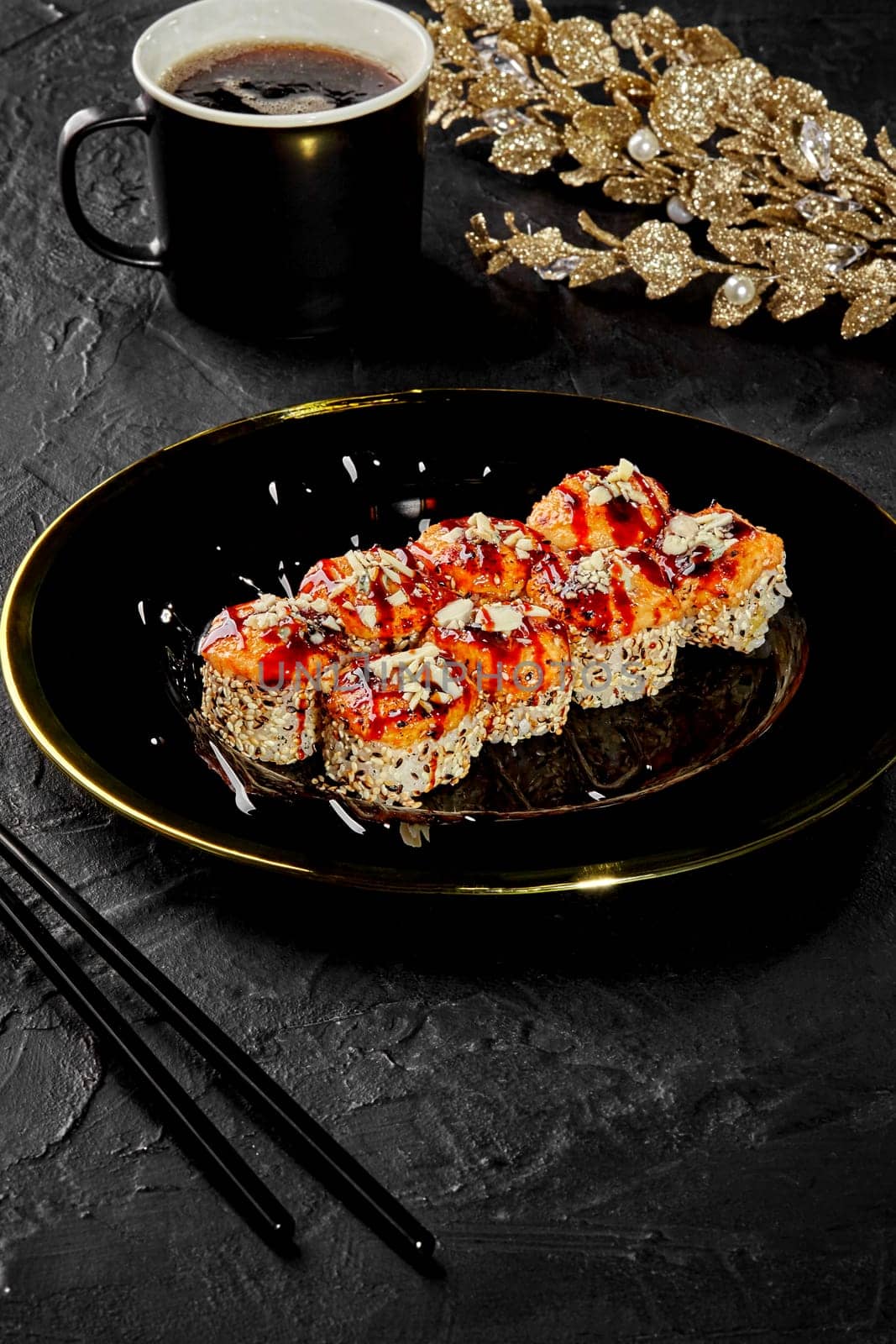 Baked sushi rolls in sesame drizzled with unagi sauce garnished with Parmesan crumbs served on black platter against dark stone surface, adorned with golden decor