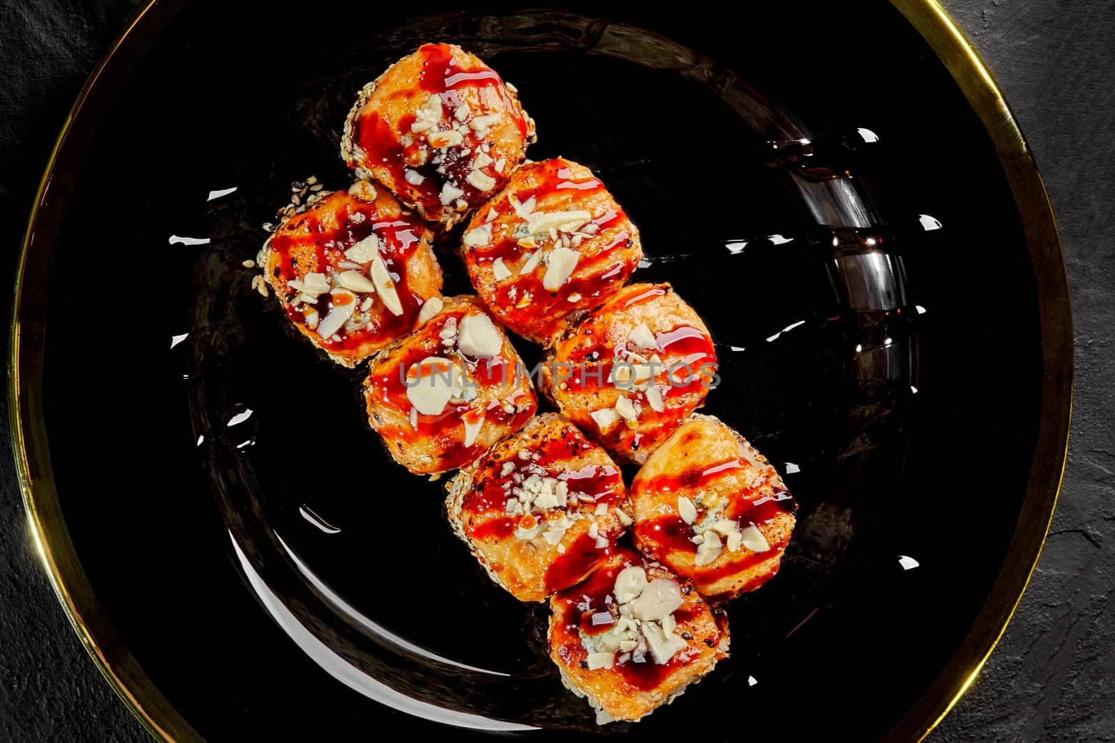 Top view of browned baked cheese caps of appetizing sushi rolls drizzled with tangy uramaki sauce and sprinkled with Parmesan crumbs, served on black and gold plate
