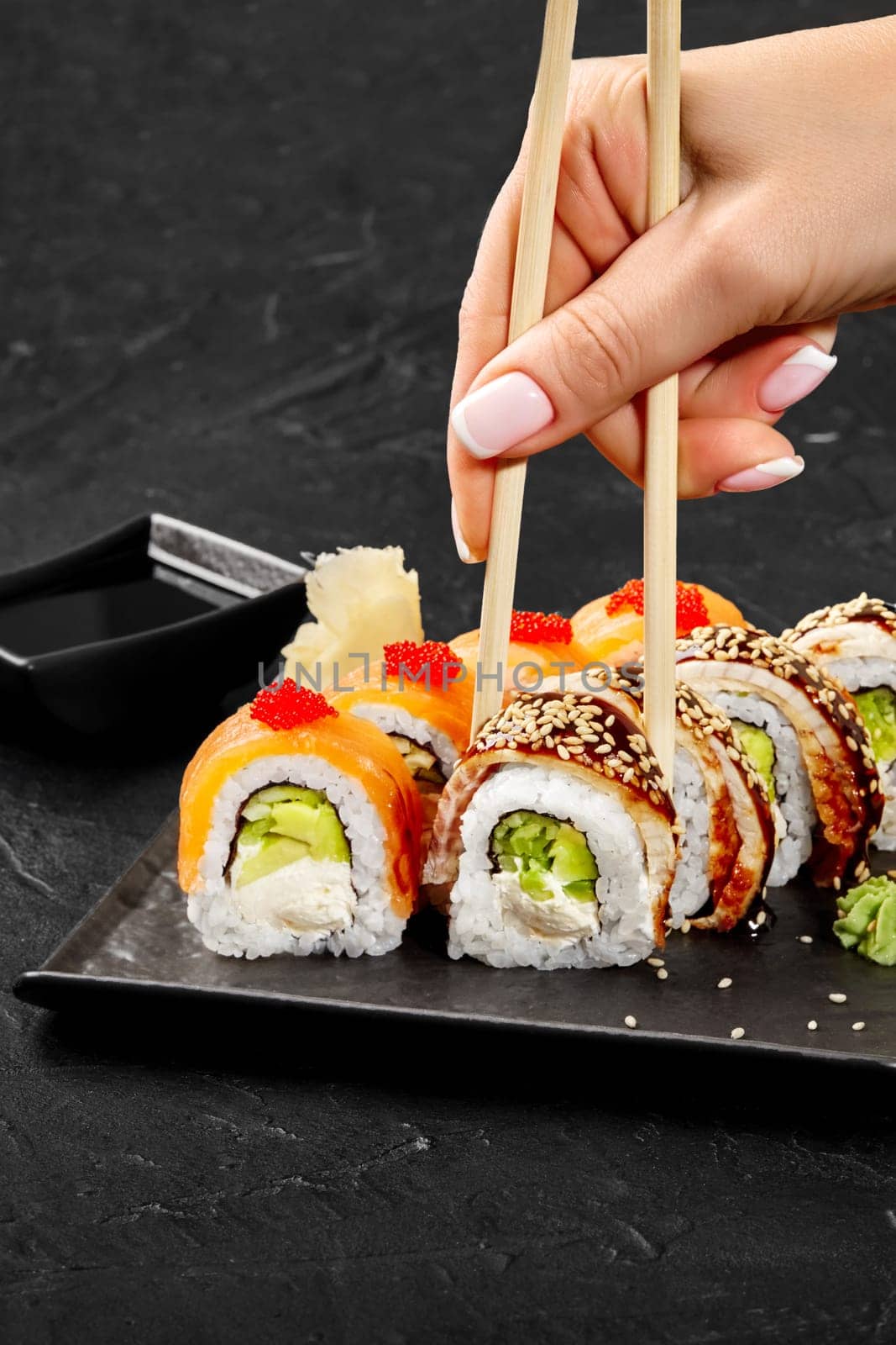 Female hand using wooden chopsticks to pick up appetizing sushi roll filled with cucumber, avocado and cream cheese topped with eel, sesame and unagi sauce from plate against black background