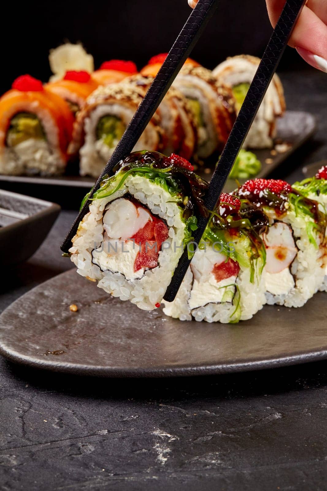 Closeup of appetizing colorful sushi roll filled with shrimp, cream cheese and tomato topped with green wakame seaweed, red tobiko roe and unagi sauce, picked up by chopsticks from black slate plate