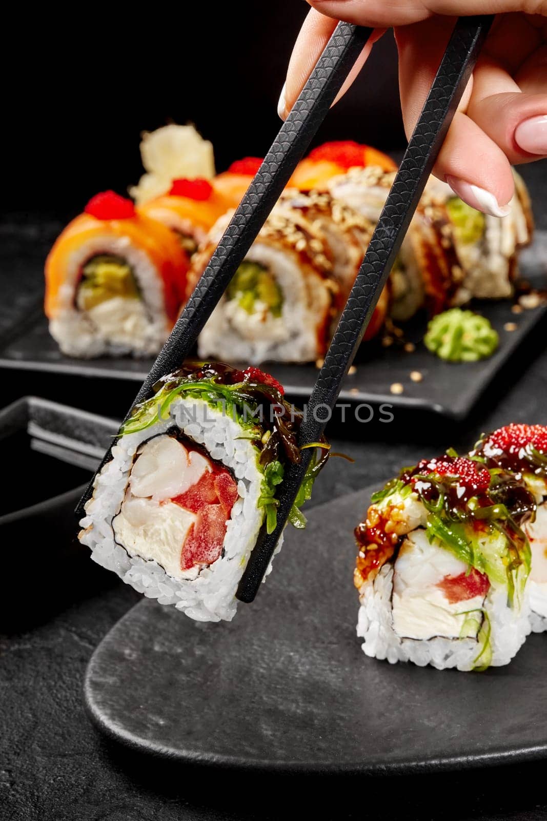 Female hand taking delicious fresh sushi roll filled with cream cheese, crab meat and tomato garnished with wakame, masago and unagi sauce, from slate plate by chopsticks. Popular Japanese snack