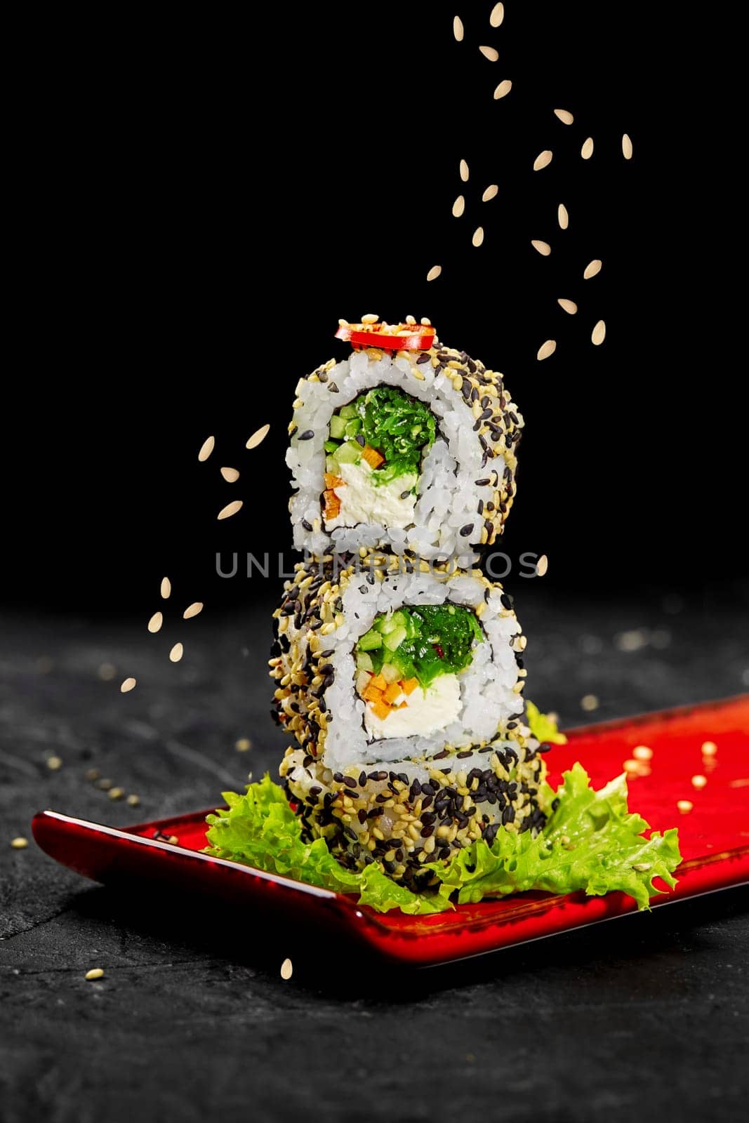 Stack of light vegetarian sushi rolls with cream cheese, carrots, hiyashi wakame, cucumber and chili pepper sprinkled by sesame seeds served on fresh lettuce leaf on vibrant red plate against black background