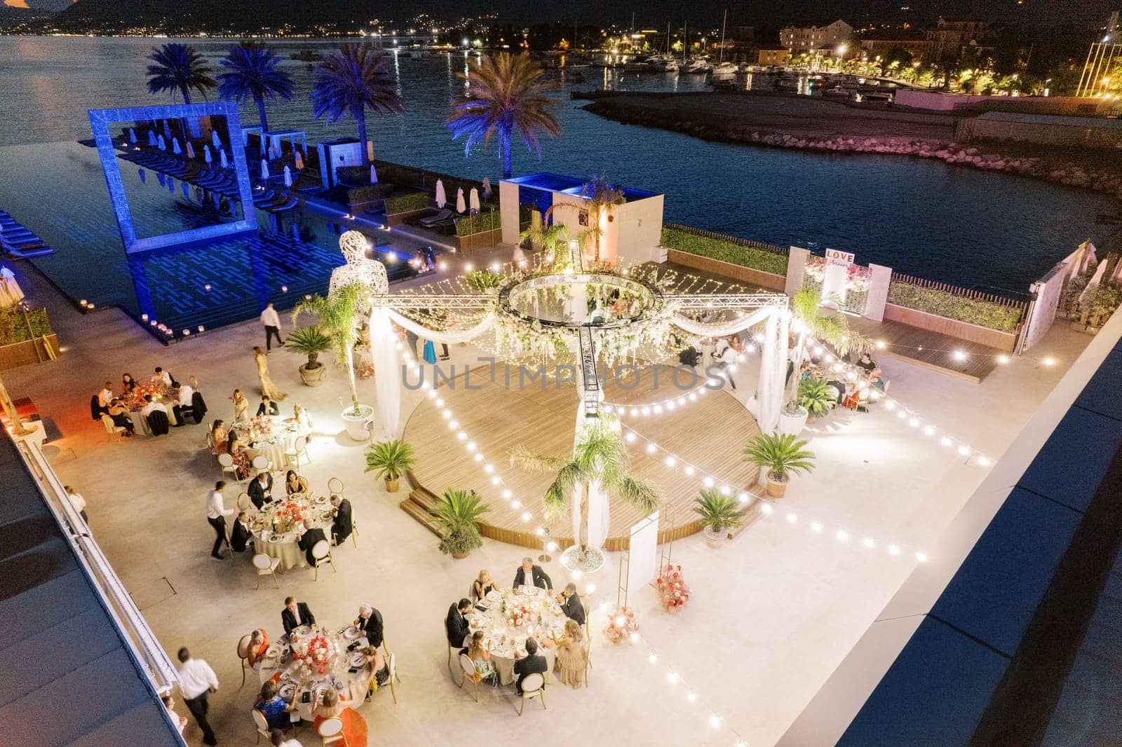 Guests sit at round tables on the pier near a stage with festive lighting and floral decorations in front of a long pool. Drone by Nadtochiy