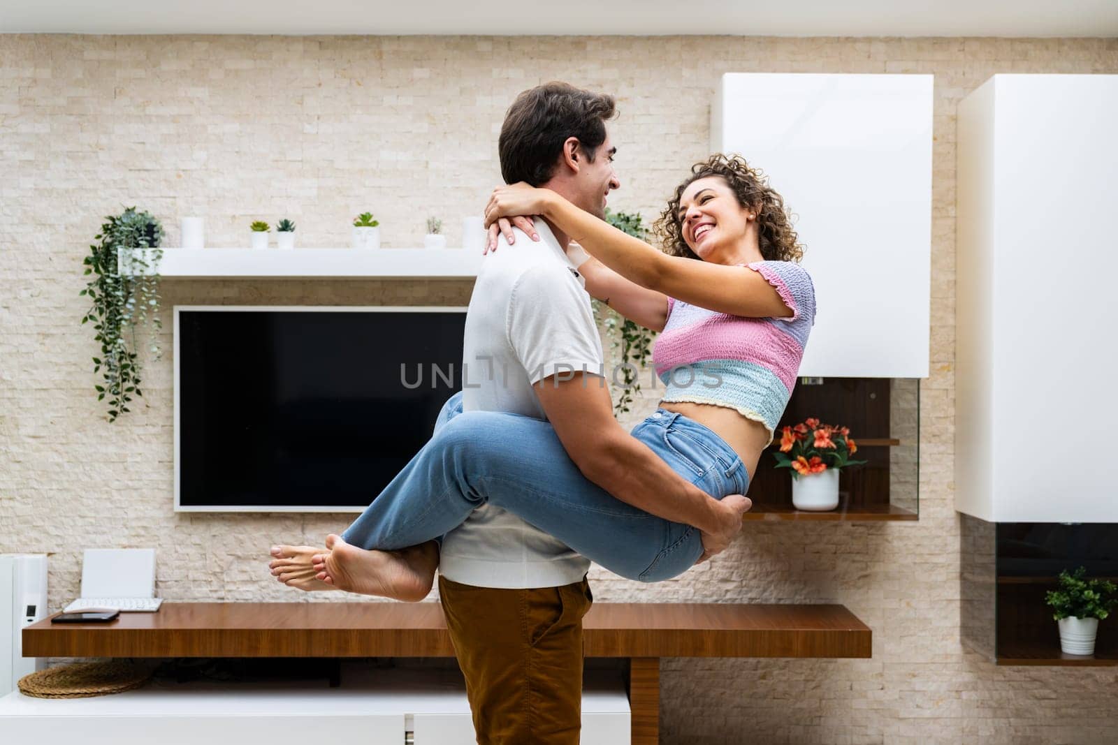 Side view of cheerful young couple hugging and looking at each other while smiling in modern living house with potted plants near tv on wall