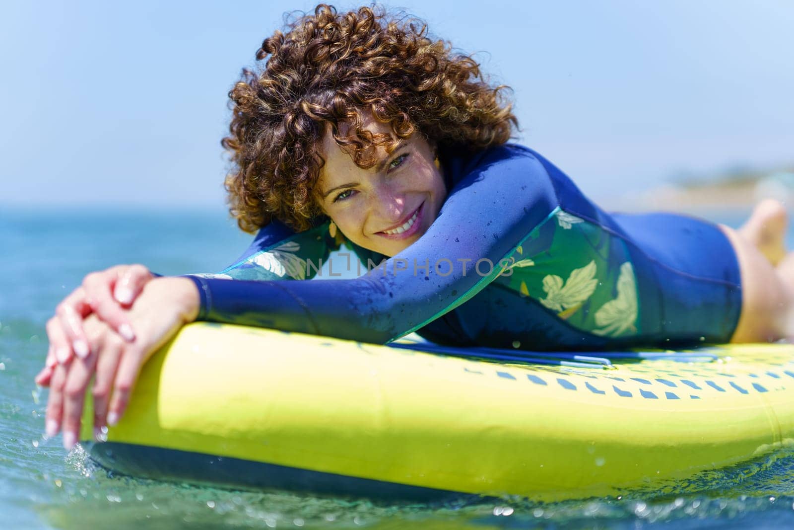 Side view of cheerful female surfer in swimwear with curly hair lying on SUP board floating on sea on bright summer day