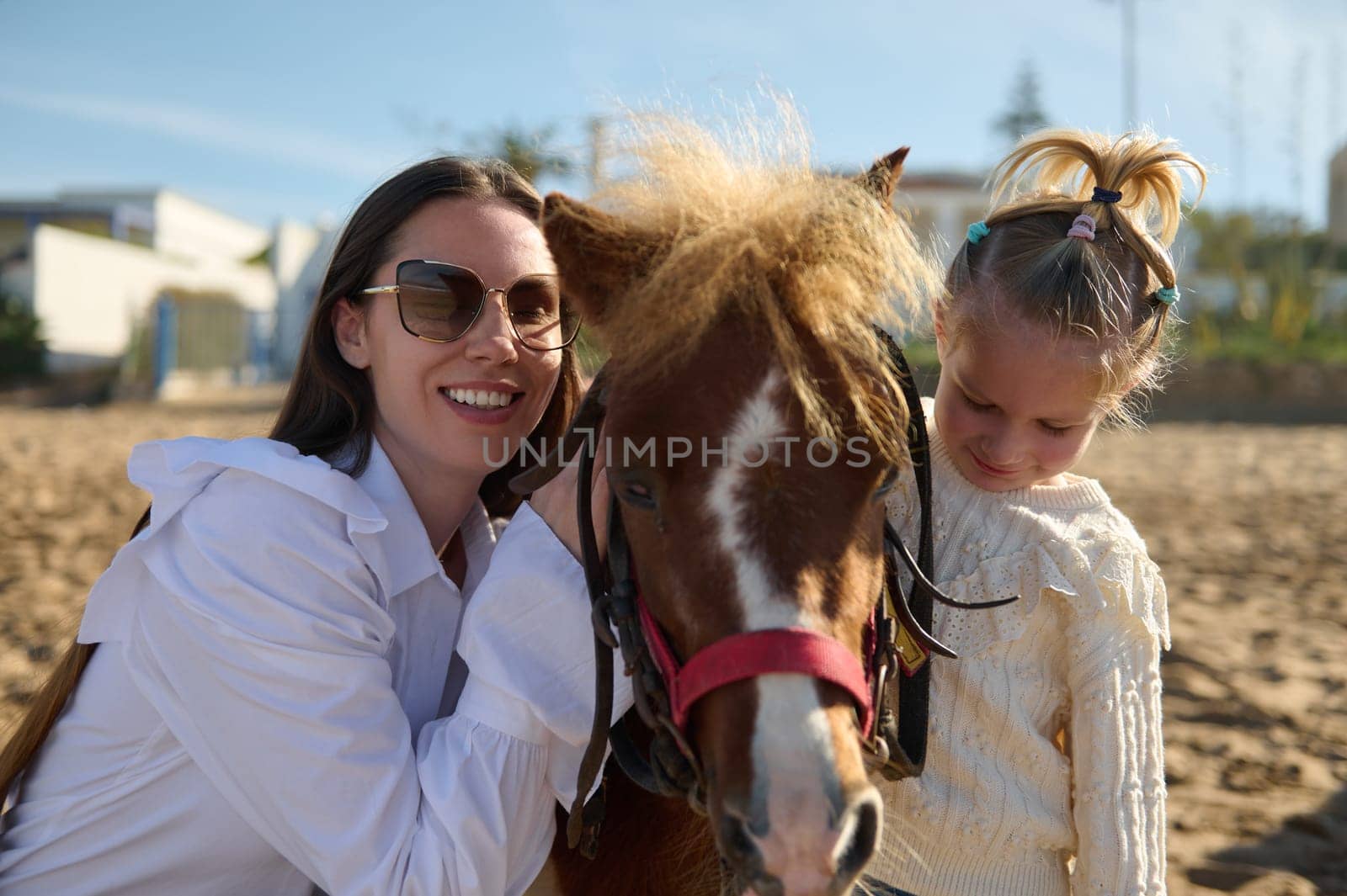 Happy young mother and daughter smiling looking at camera, hugging and posing with a little pony horse. People. Leisure and hobbies. Active healthy lifestyle