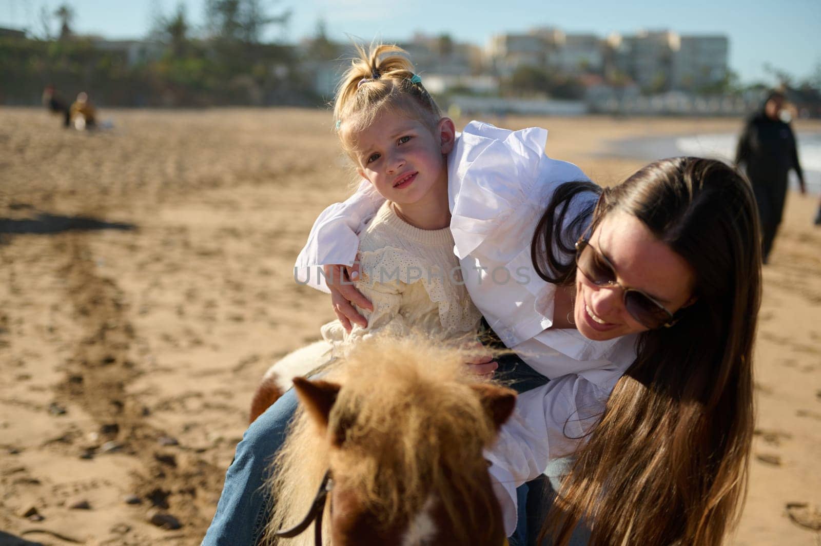 Smiling mother stroking a little pony while supports her daughter on learning horse riding on the sandy beach. People. Active healthy lifestyle. Hobbies and leisure